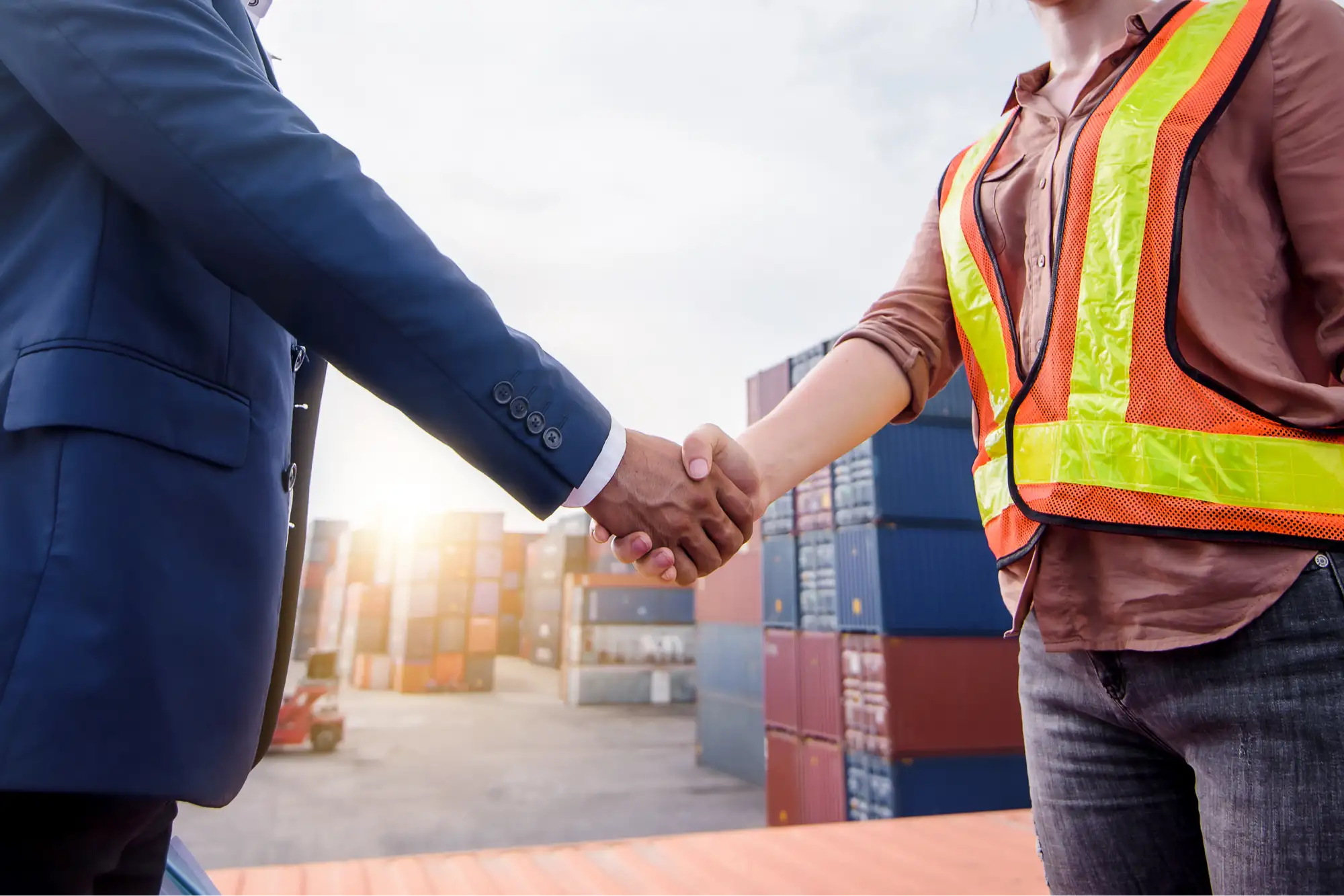 A business partner and a site worker shaking hands in front of stacked shipping containers